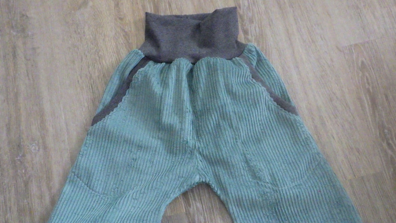 Puede incluir: Un pantal&oacute;n de pana azul verdoso con una cintura de canal&eacute; gris. El pantal&oacute;n tiene dos bolsillos grandes en la parte delantera.