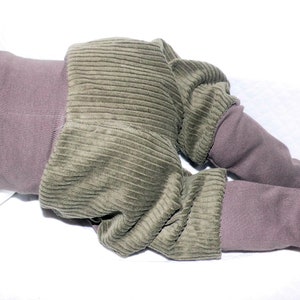 May include: Olive green corduroy baby trousers with brown ribbed cuffs. The trousers are folded and laid on a white surface.