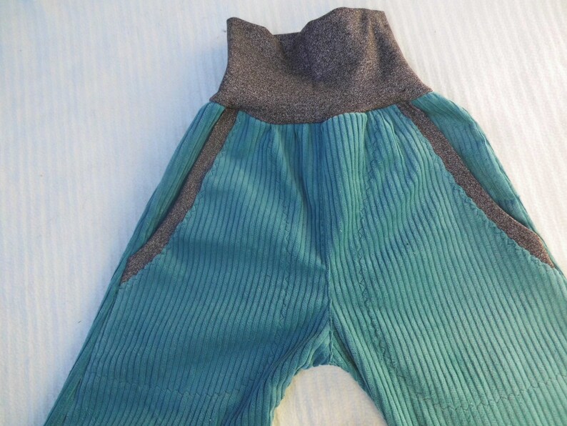 Puede incluir: Pantal&oacute;n de pana color turquesa con cintura el&aacute;stica gris oscuro y ribetes negros a los lados.