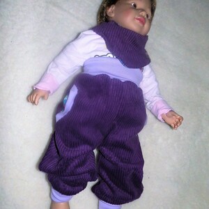Op de afbeelding: Een baby-outfit van paarse corduroy met een lichtpaarse en witte top en een roze en witte slabbetje.