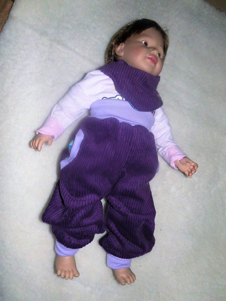 Op de afbeelding: Een baby-outfit van paarse corduroy met een lichtpaarse en witte top en een roze en witte slabbetje.