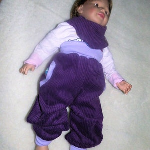 Op de afbeelding: Een babypop-outfit van paarse corduroy met een lavendelkleurig topje en een wit shirt met lange mouwen eronder. De pop heeft bruin haar en draagt een paarse nekwarmer.