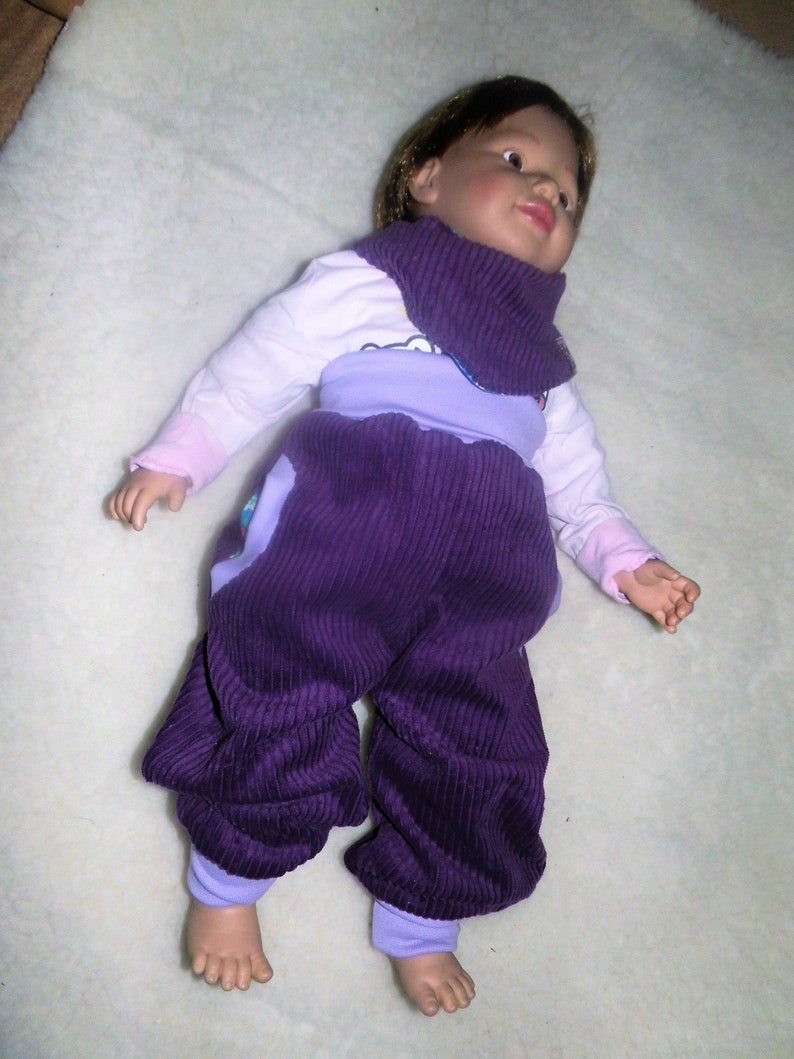 Op de afbeelding: Een babypop-outfit van paarse corduroy met een lavendelkleurig topje en een wit shirt met lange mouwen eronder. De pop heeft bruin haar en draagt een paarse nekwarmer.