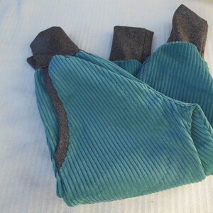 Puede incluir: Un pantal&oacute;n de pana azul turquesa con pu&ntilde;os de canal&eacute; grises. El pantal&oacute;n est&aacute; doblado por la mitad y colocado plano sobre una superficie blanca.
