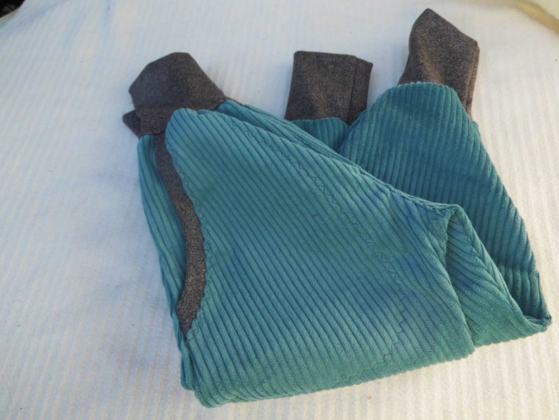 Puede incluir: Un pantal&oacute;n de pana azul turquesa con pu&ntilde;os de canal&eacute; grises. El pantal&oacute;n est&aacute; doblado por la mitad y colocado plano sobre una superficie blanca.