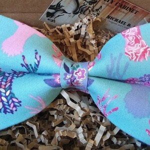 May include: A turquoise bow tie with a repeating pattern of deer silhouettes in pink, purple, and white. The bow tie is made of fabric and is presented in a brown box with paper filler.