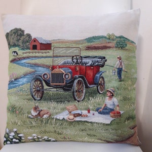 May include: A vintage red car with a white steering wheel and black tires is parked in a field. A person is sitting on the grass in front of the car, and a dog is lying on the grass behind the car.