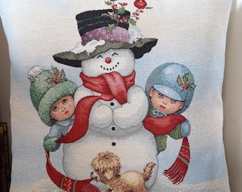 Op de afbeelding: Een decoratief kussen met een wintertafereel. Het toont een sneeuwman met een hoge hoed en rode sjaal, omarmd door twee kinderen met mutsen en sjaals. Een kleine hond staat aan de voet van de sneeuwman. De achtergrond is lichtblauw.