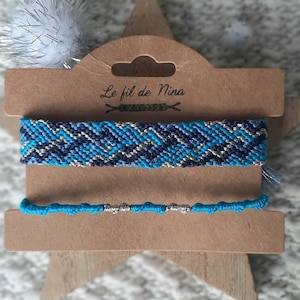 Puede incluir: Dos pulseras de la amistad azules exhibidas en un soporte de cartón marrón en forma de estrella. La pulsera superior tiene un patrón de chevrón en tonos azules y plateados. La pulsera inferior es de color turquesa con cuentas plateadas. El texto "Le fil de Nina" es visible.