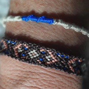 Può includere: Sono mostrati due braccialetti fatti a mano. Il braccialetto superiore è bianco con un dettaglio blu. Il braccialetto inferiore è nero con un motivo floreale rosa, bianco e blu. I braccialetti sono fatti di fili intrecciati.