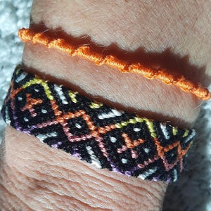 Peut inclure: Deux bracelets sont présentés. Le premier est un bracelet d'amitié tissé avec un fond noir et des motifs géométriques orange, jaune, blanc et violet. Le second est un simple bracelet en cordon torsadé orange.