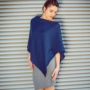 Könnte beinhalten: Eine Frau in einem marineblauen Poncho und einem grauen Rock, die vor einer silbernen Metallwand steht. Sie trägt braune Schuhe mit Absatz.