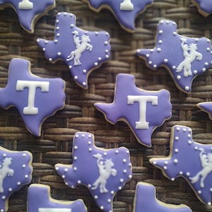 May include: Purple and white Texas-shaped cookies decorated with a white 'T' and a white silhouette of a cowboy riding a horse.