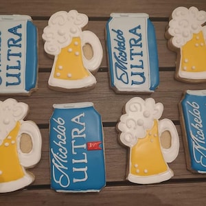 May include: Six decorated sugar cookies, four are shaped like beer mugs with white foam and yellow beer, and two are shaped like a can of Michelob Ultra beer with blue and red accents.