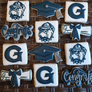 May include: A set of 10 decorated sugar cookies featuring a graduation cap, a diploma, the word "Congrats", and a bulldog mascot with the letter "G" on its chest. The cookies are white with blue and black icing.