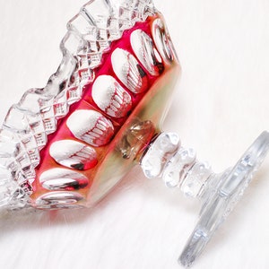 May include: A clear glass footed bowl with a ruffled rim and a red interior. The bowl has a decorative pattern on the rim and the interior is decorated with white oval shapes. The stem is clear glass.