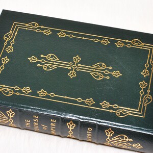 May include: A hardcover book titled "The Course of Empire" by DeVoto. The book has a dark green cover with gold decorative accents and gold lettering. The book is closed and resting on a white surface.