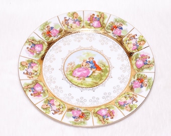 May include: A decorative white porcelain plate with a gold rim and a central image of a couple in a garden. The plate features a repeating pattern of couples in period clothing, with pink and blue accents.