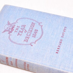 May include: A light blue hardcover book titled "The Year of Decision 1846" by Bernard DeVoto. The title and author's name are in burgundy, with an eagle emblem above the title. The book has a textured cover.