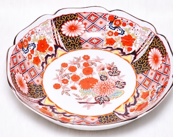 May include: A decorative, scalloped-edge ceramic bowl with a white interior and a detailed floral and geometric pattern. The design features red, orange, and blue flowers, with black and gold accents. The bowl has a brown rim.