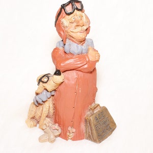 May include: A whimsical figurine of an elderly man in a pilot's helmet and goggles, with a dog wearing glasses. The man is in a red coat, and the dog is tan. A small suitcase with the text "Ace Flying School" is at the man's feet.