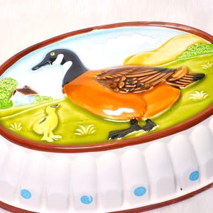 May include: Oval ceramic serving dish with a goose and chick design. The dish features a white base with blue accents and a brown rim. The goose has orange and brown feathers, a black head, and is set against a green field with a small house.