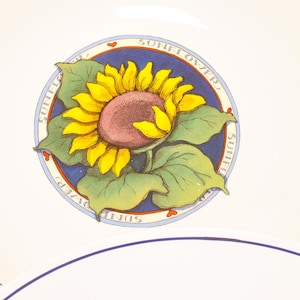 May include: White ceramic plate with a blue rim and a central sunflower design. The sunflower has yellow petals, a maroon centre, and green leaves. The words "SUNFLOWER" are written around the flower.