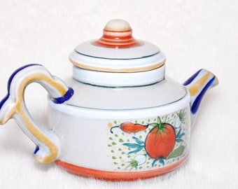 May include: A ceramic teapot with a white base, decorated with a red tomato and chili pepper design. The handle and spout have blue and yellow stripes. The lid is white with red and orange accents.