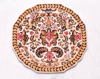 May include: A decorative plate with a scalloped edge and a detailed floral pattern. The plate features a cream-colored background with a repeating design of red, brown, and black floral motifs. The border is a warm tan color with a leaf design.