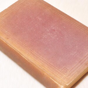 May include: A vintage book with a faded, textured cover. The cover is a muted reddish-pink color with a rectangular border design. The spine and edges of the book are a warm, light brown.