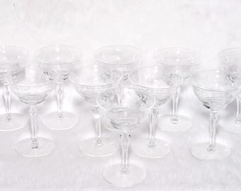May include: A collection of clear, stemmed champagne glasses. Each glass features a round bowl with etched floral designs and a slender stem. The glasses are arranged on a white surface, suggesting a formal setting or event.