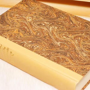 May include: A hardcover book with a marbled brown and gold cover. The spine is a light tan color with gold lettering. The book is open, revealing the pages. The book is resting on a white surface.
