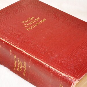 May include: A close-up of a vintage, red leather-bound book titled "The New Century Dictionary." The cover features embossed decorative patterns and gold lettering. The book is Volume Two, with "pock-marks, zymurg & supplements" visible.