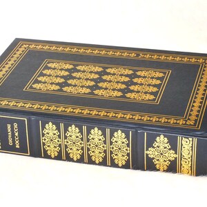 May include: A hardcover book titled "The Decameron" by Giovanni Boccaccio. The book has a dark blue cover with gold decorative patterns and text. The edges and spine are also adorned with gold designs. The book is lying on a light-colored surface.
