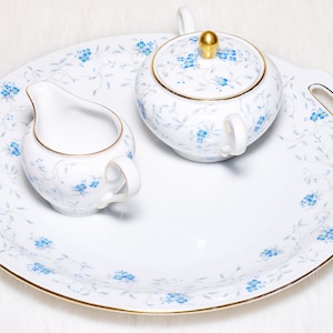 May include: A white porcelain tea set with a floral pattern. The set includes a serving tray, a creamer, and a sugar bowl with a lid. The floral design features blue flowers and gold trim.