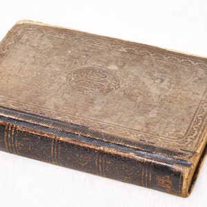 May include: An antique book with a worn, brown leather cover. The cover features a decorative border and a central embossed design. The spine is dark brown with gold lettering. The book appears aged, with signs of wear.