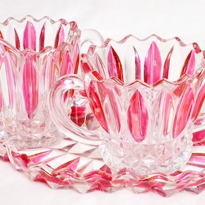 May include: A clear glass creamer and sugar bowl set with a matching tray. The set features a pink and clear design with a scalloped rim and diamond-shaped cuts. The creamer has a handle and spout.