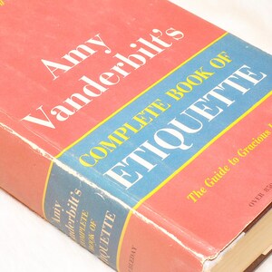 May include: A vintage hardcover book titled "Amy Vanderbilt's Complete Book of Etiquette." The book has a red cover with a blue and yellow banner. The title and author's name are in white. The book is a guide to gracious living.