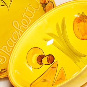 May include: Two yellow ceramic spaghetti bowls with hand-painted illustrations of tomatoes, onions, and cheese. One bowl has the word "Spaghetti" written in cursive. The bowls are oval-shaped and have a glossy finish.