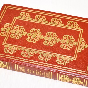 May include: A hardcover book with a red leather cover and gold filigree design. The book's cover features a rectangular gold frame with ornate gold embellishments. The spine displays the title and author in gold lettering.