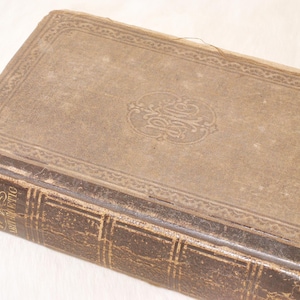 May include: Antique book with a worn, brown leather cover. The cover features an embossed decorative border and a central design. The spine is also leather, with gold lettering. The book's edges show signs of age.