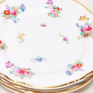 May include: A stack of white porcelain plates with a scalloped gold rim. Each plate is decorated with colorful floral bouquets in red, pink, blue, and yellow. The plates are stacked on top of each other.