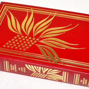 May include: A red, hardcover book with gold foil accents. The cover features a stylized design of a burst with stars, and the spine displays the title "No Safe Place" and author's name. The book is a signed first edition.