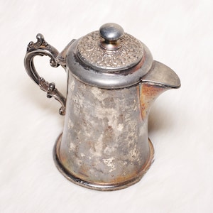 May include: Antique silver-plated coffee pot with ornate handle and lid. The pot has a spout for pouring and a decorative floral pattern on the lid. The silver finish shows signs of age and use, adding to its vintage charm.