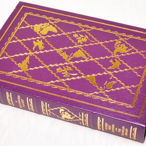 May include: A hardcover book titled "A Patchwork Planet" by Anne Tyler. The book has a deep purple cover with gold embossed designs and text. The cover features a grid pattern with various gold illustrations. The spine reads "Signed First Edition The Franklin Library."