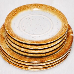 May include: A stack of six ceramic saucers with a speckled white center and a brown, textured rim. The saucers are stacked on top of each other, showcasing their design. The plates are suitable for a kitchen or dining setting.
