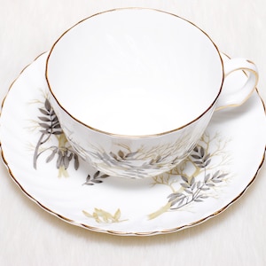 May include: White teacup and saucer set with a gold rim and a decorative tree branch design in gray and gold. The teacup has a handle and sits on a matching saucer.