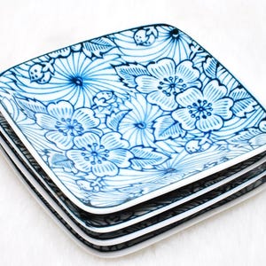 May include: A stack of four square plates with a white and blue floral design. The plates feature a detailed pattern of flowers and leaves in shades of blue against a white background. The plates have a white border.