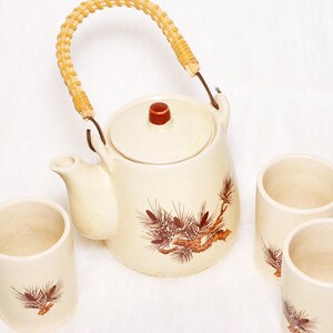 May include: Cream ceramic teapot with a woven handle and a red knob, alongside three matching cups. Each item has a brown and red botanical design. The set is suitable for serving tea.