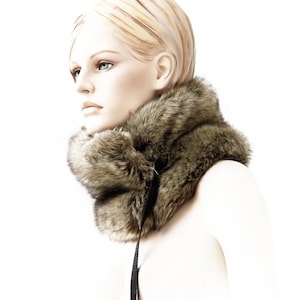 May include: A brown faux fur neck warmer with a black adjustable strap. The neck warmer is worn by a mannequin with blonde hair.